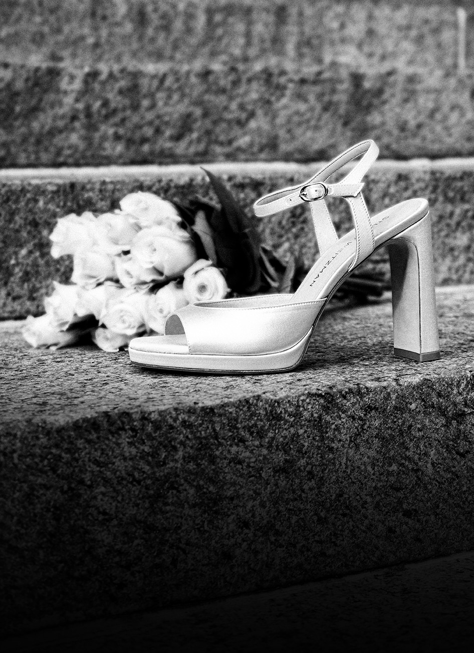 Black and white photo of a white platform sandal with ankle strap and block heel displayed with wedding flowers.