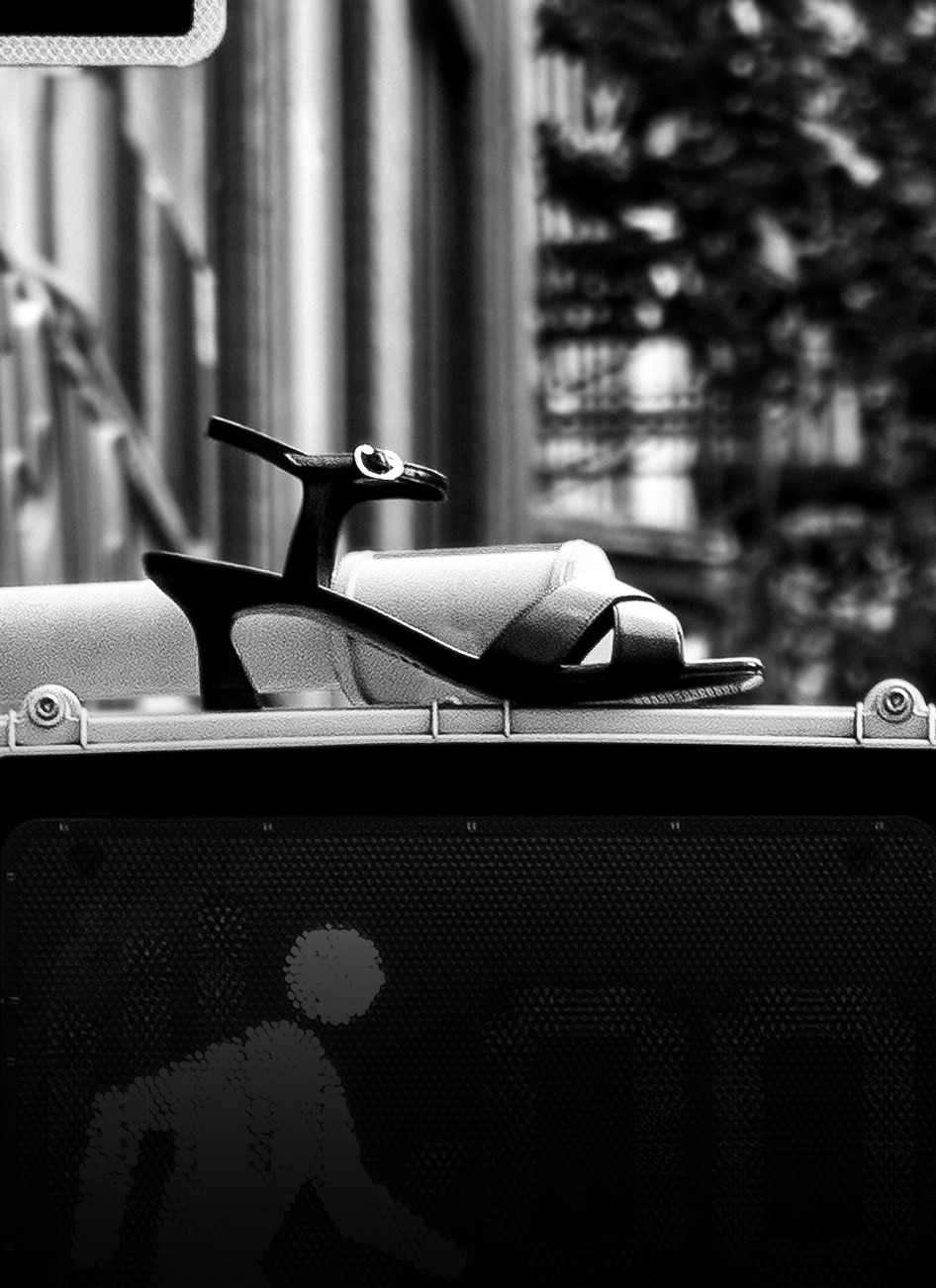 Black and white photo of a woman's black ankle strap sandal with kitten heel displayed on a platform.