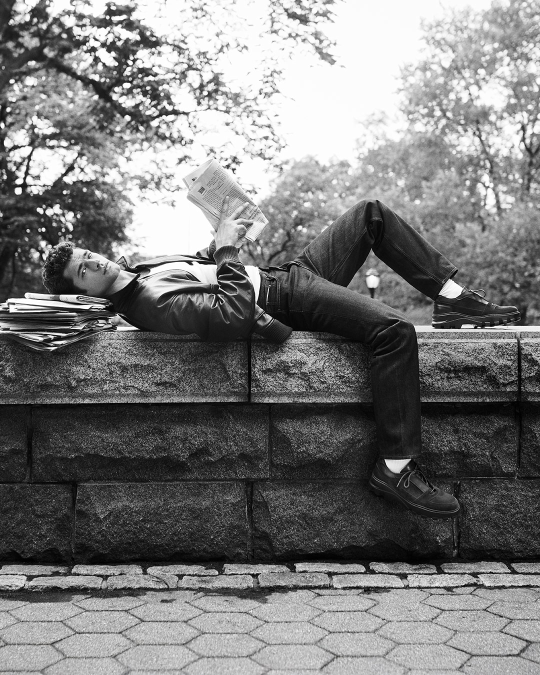Man relaxing on a stone wall, holding a newspaper, and wearing suede and leather panel sneakers with lug soles.